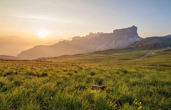 A scenic landscape of rolling green fields leading to rugged mountains at sunrise, with soft light illuminating the valley and partly cloudy sky.