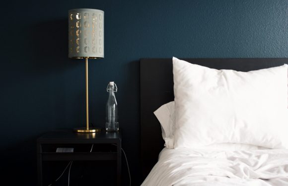 A nightstand with a glass bottle, a gold desk lamp with a patterned shade, and a power outlet visible, next to a bed with white pillows and bedding against a dark wall.