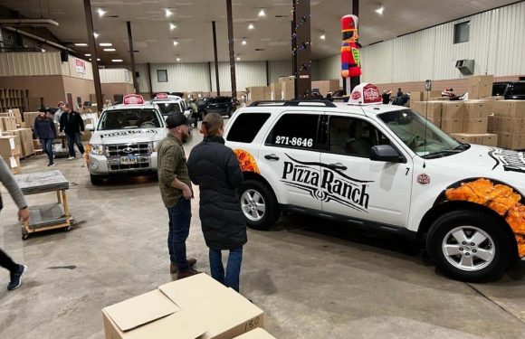 A Pizza Ranch vehicle is parked inside a warehouse-like space with people gathering around. There are large stacks of cardboard boxes and a decorative nutcracker and nutcracker figure in the background.
