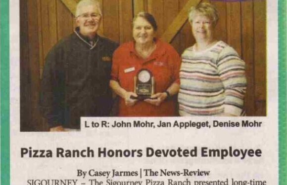 A woman holding a plaque is flanked by a man and a woman, standing in front of a wooden backdrop. The woman wears a red shirt, the man a dark jacket, and the other woman a striped top. The image appears to be a newspaper clipping celebrating an employee's 25 years of service.