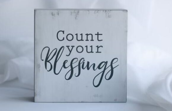 A square wooden sign with a distressed white finish displaying the words "Count your blessings" in black, mixed fonts on a soft white background.