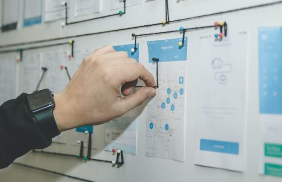 A person pins a map on a bulletin board. The individual wears a black jacket with a smartwatch on their wrist, and the board has multiple pinned papers and notes.