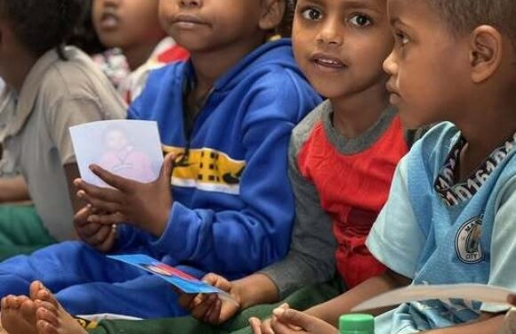 Group of children sitting together indoors on a colorful mat, holding papers and a bottle of beverage, with their legs crossed and attentive expressions.