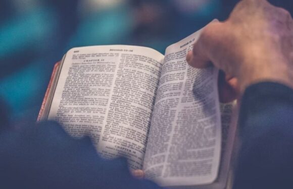 A person holding an open Bible, with focused fingers on the right page, displaying text from the book of Jeremiah. The background is blurred with shades of blue.