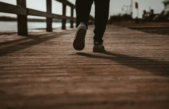 Person walking on a wooden dock near water, wearing black pants and sneakers, with an overcast sky and distant buildings in the background.