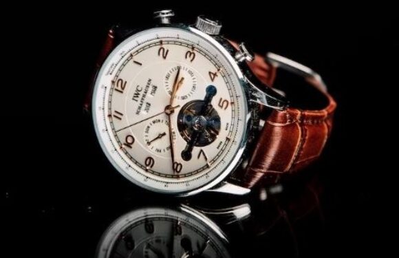 A luxury wristwatch with a white dial, multiple subdials, and a brown leather strap, displayed against a black background with a reflective surface.