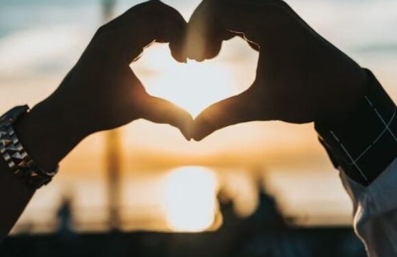 Two hands form a heart shape with the sun shining through, set against a blurred background of a sunset over a body of water.