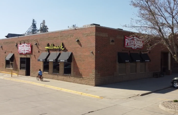 A person riding a bike in front of a large Pizza Ranch restaurant with "Fallmark" signage, brick exterior, and black awnings; leafless tree and parked car nearby.