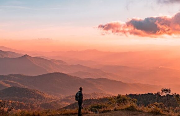 A person with a backpack standing on a hilltop, overlooking layered mountains during a colorful sunset with clouds and warm orange-pink hues.