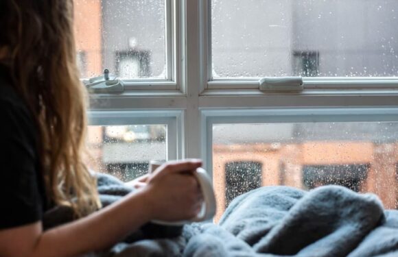 A person with curly hair sits by a rainy window, holding a mug and wrapped in a blanket, with raindrops on the window and blurred buildings outside.