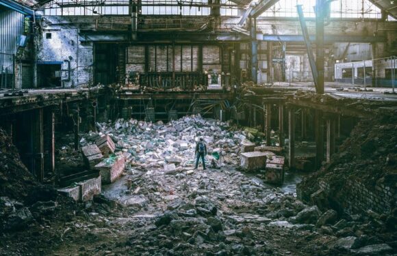 A person wearing a blue jacket and jeans stands amid the rubble and debris inside a large, abandoned industrial building with exposed brick and metal structures.