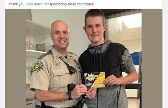 A young man in a gray and black hoodie holding a certificate stands next to a smiling police officer in uniform, in an indoor hallway with shelves in the background.