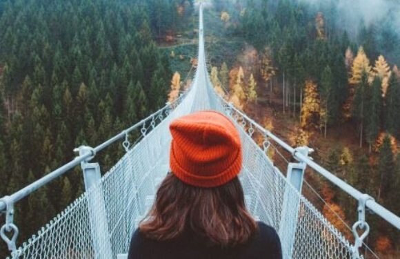 Person with brown hair and red beanie standing on a suspension bridge overlooking a forested landscape with misty hills and trees in autumn colors.