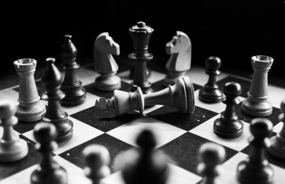 A chessboard with black and white pieces, including a toppled king, set against a dark background, with shadows and highlights emphasizing the pieces.