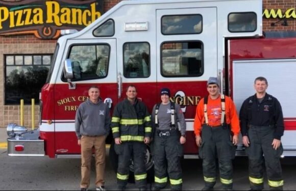 Five firefighters stand in front of a red fire truck with a Pizza Ranch sign in the background, all wearing their gear and uniforms.