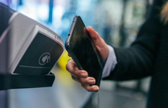 A person in a black suit is scanning their phone at a contactless payment terminal in a public space with a blurred background.
