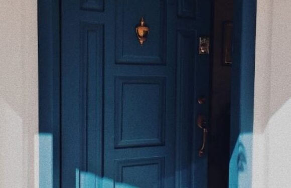 A blue arched door set in a beige wall, with a gold door knocker and a peephole, partially open to reveal a dark interior.