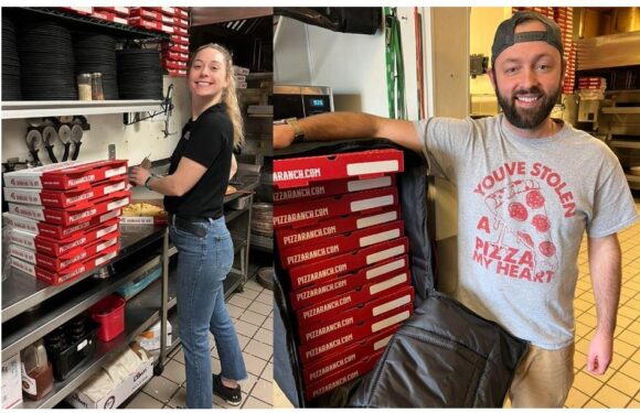 Two employees in black and gray Pizza Ranch shirts prepare and deliver pizzas in a kitchen, with stacks of pizza boxes and kitchen shelves visible.
