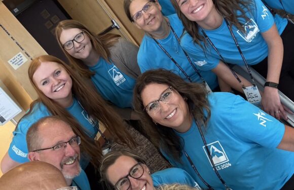 Group of smiling people wearing blue shirts, some with glasses, gathered indoors near a wooden wall with a sign, posing for a photo.