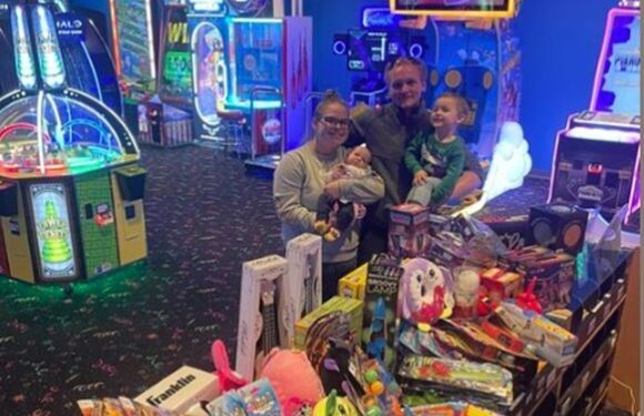Three children, two girls and a boy, with the girl holding a baby, are smiling and sitting at a table filled with toys in an arcade or entertainment center.