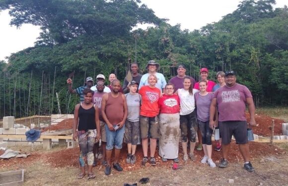 Group of people standing outdoors at a construction site with trees in the background, some in casual clothing and others in work attire.