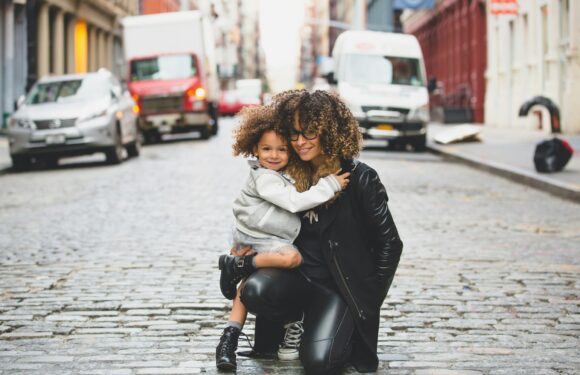 A woman with curly hair and glasses kneels on a cobblestone street, holding a smiling young girl with curly hair; parked cars and buildings are visible in the background.