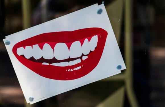 A sign with a large, open-mouthed smile showing teeth, attached with four corner fasteners, mounted on a transparent surface.