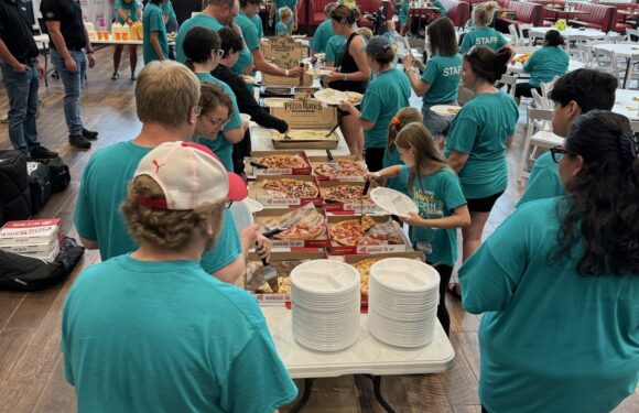 People in turquoise staff shirts and children are serving and eating pizza at a buffet table during an event in a bright, spacious room with large windows.