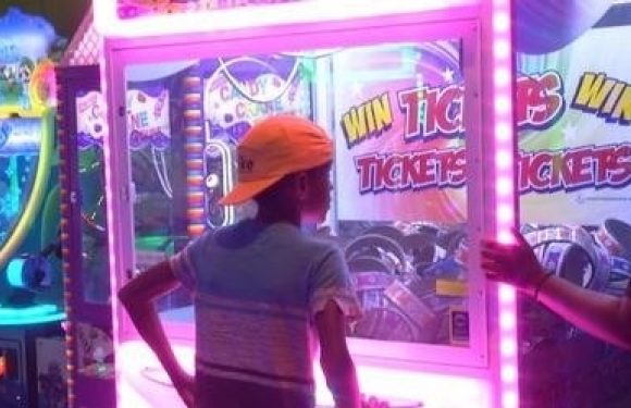 A child in an orange cap and striped shirt plays a game at an arcade, with a brightly lit ticket redemption machine behind them.