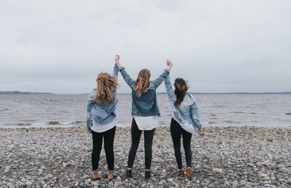Three women standing on a rocky beach, holding hands and raising their arms in celebration against a cloudy sky and calm water. They are dressed in denim jackets and black pants.