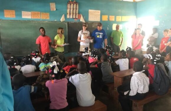 Children sitting at desks in a classroom, with several adults standing at the front of the room. The room has a dark green chalkboard and educational posters on the walls.