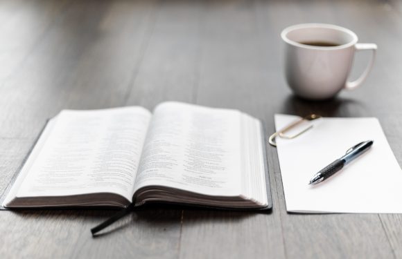 Open Bible, blank sheet of paper with a pen, look like coffee mug, all on a wooden surface.