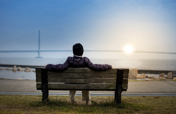 A person in a dark jacket sits on a wooden bench facing the ocean at sunset, with a bridge visible in the background over calm water.