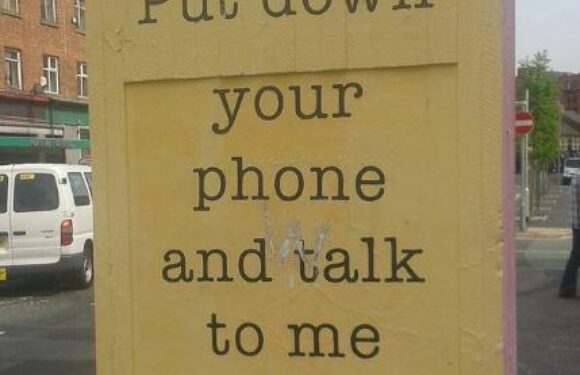 A yellow poster with black text on a city street corner, instructing to put down your phone and talk, signed by Micah Purnell. The background shows parked cars and buildings, some with signs including "Habib".