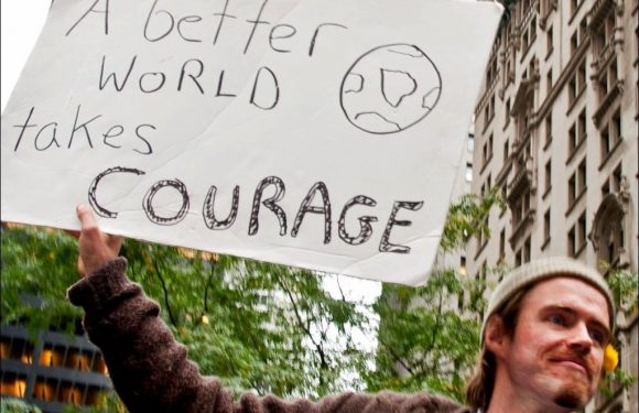A person holds a sign that reads "A better world takes COURAGE" with a drawing of Earth, standing in front of tall buildings and trees during the day.