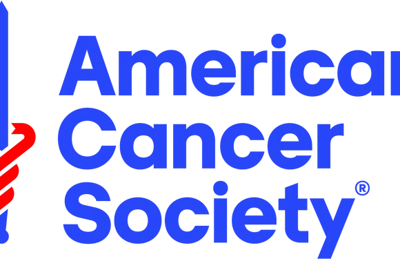 A person wearing a red and white striped apron and cap stands next to a rotisserie with various meats, holding a tray, in front of a blue and white sign for a cancer society.