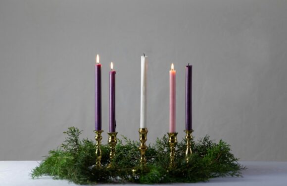 An Advent wreath with five lit candles in purple, pink, and white, arranged in gold holders, surrounded by green foliage, on a white surface with a plain gray background.