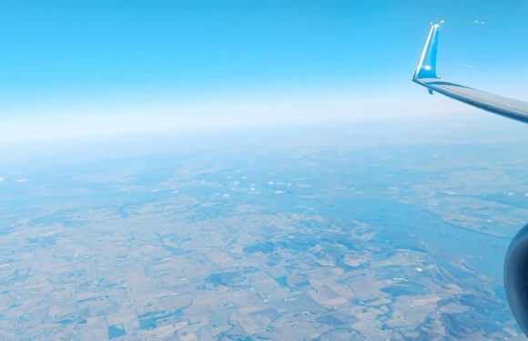 View of farmland terrain below from an airplane window, with a blue sky and the airplane's wingtip visible on the right.