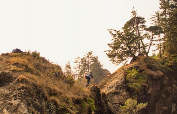 A person climbing or scrambling on rugged, rocky terrain surrounded by trees, with a backpack resting on the ground nearby.