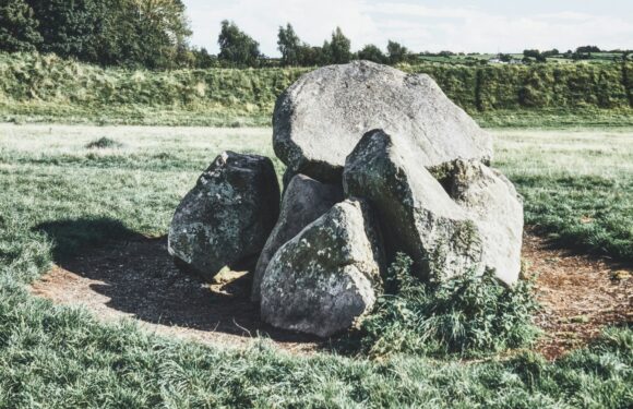 A large group of gray rocks arranged in a circle on a grassy field, with trees in the distance and a partly cloudy sky overhead.