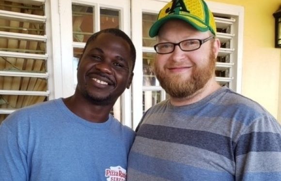 Two smiling men, one in a blue Pizza Ranch T-shirt and the other in a striped shirt and green baseball cap, stand indoors near a window with white blinds.