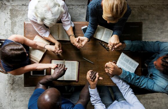 Six diverse people sit around a wooden table, holding hands, with open books and a smartphone placed on the table. They are engaging in a group prayer or support.