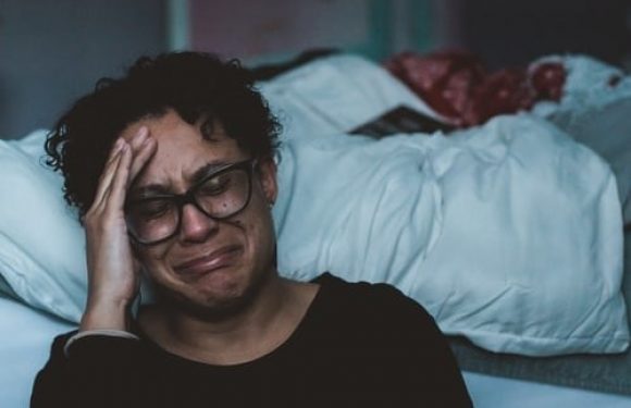 A person with glasses sitting on a bed, holding their head and appearing distressed or upset, in a dimly lit room with blankets and pillows.