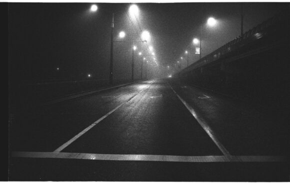 Empty, wet road at night with streetlights and power lines, foggy atmosphere, and no visible vehicles or pedestrians.