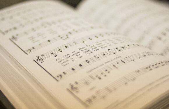 An open hymn book on a music stand showing sheet music and lyrics for a song titled "An-cient" with musical notation and printed words.