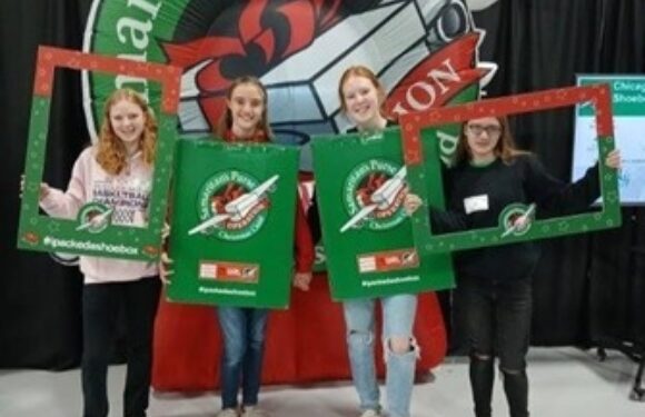 Four young women stand together holding pizza boxes and red photo props, smiling in front of a large Pizza Ranch promotional backdrop.