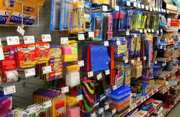 A retail shelf displays various school and office supplies, including colorful pens, glue sticks, notebooks, and binders organized on hooks and shelves.
