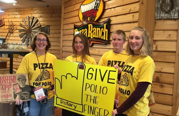 Four teenagers wearing yellow "Pizza Polio" t-shirts stand inside a wooden-paneled restaurant, holding a yellow sign that says "GIVE POLIO THE FINGER" with a hand showing the middle finger.