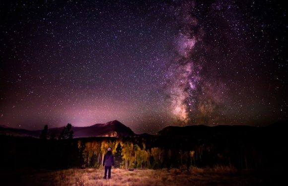 A person wearing a Pizza Ranch uniform stands in a field at night, gazing up at a star-filled sky and the bright, colorful Milky Way galaxy above mountainous landscape.