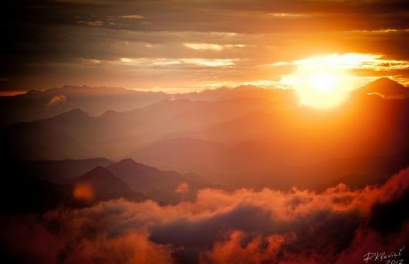 A vibrant sunset over layered mountains with clouds illuminated in shades of orange and pink, casting a warm glow across the landscape.